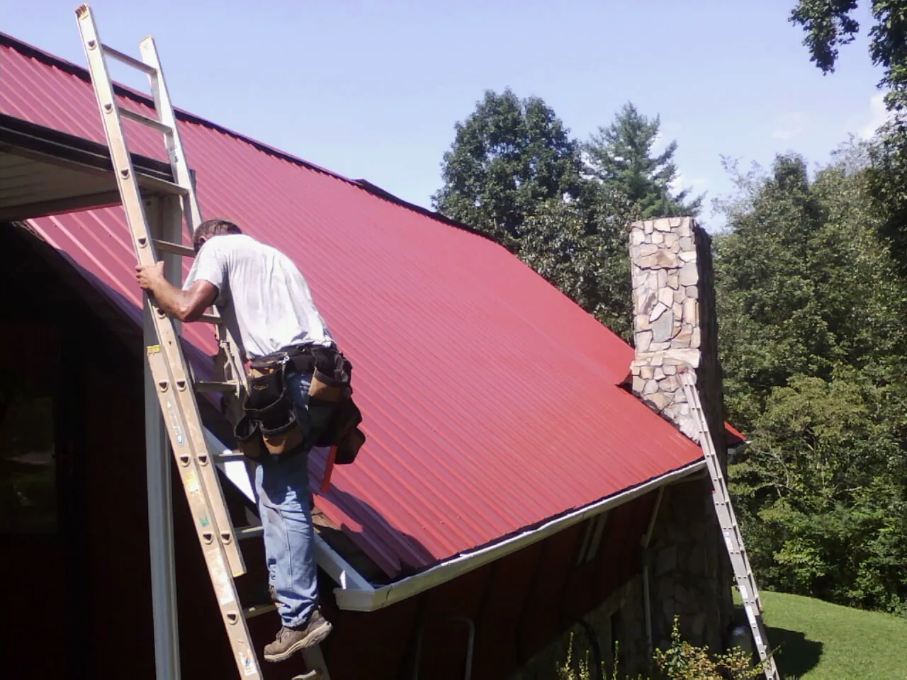 Roofer on a ladder beside a red metal roof and stone chimney — Soffit & Fascia Repair by Roofing Brothers in Wilbur